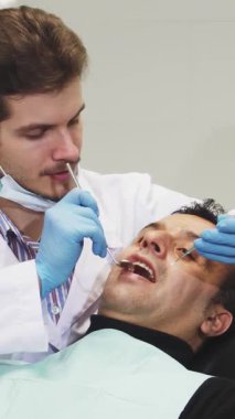 Gıcır gıcır beyaz bir ceket ve mavi eldivenler giymiş neşeli genç bir dişçi, rahat yetişkin erkek hastasının dişlerini parlak, modern bir dişçi koltuğunda incelerken kendine güvenle ışıldıyor. Parlak, steril klinik profesyonellik ve sıcaklık yayıyor.