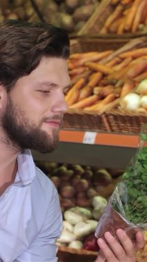 Şık sakallı genç beyaz bir adam canlı bir süpermarkette canlı bir dizi renkli ürünle çevrili bir tepside taze fesleğen kokulu bir demet koyar. Mutluluk ifadesi aşçılık tutkusunun özünü yansıtıyor.
