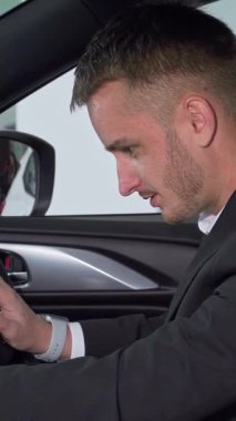 Özel dikim takım elbise giymiş sofistike bir işadamı, şık, modern bir arabanın sürücü koltuğuna güvenle oturur ve özenle son model iç hatlarını inceler. Bu dinamik sahne heyecan verici bir keşif anını yakalar.