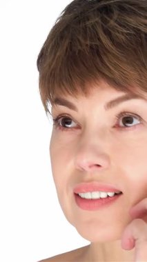 Kısa, şık saçlı, zarif orta yaşlı bir kadın, el değmemiş beyaz bir fon karşısında yanağını nazikçe okşuyor, kendine güven ve doğal güzellik saçıyor. Bu büyüleyici yakın çekim onun kusursuz cildini ve huzurlu ifadesini yakalıyor, izleyicileri davet ediyor.