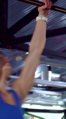 Odaklanmış genç bir kadın enerjik bir spor salonunda barfiks çekerken gücünü ve atletizmini sergiliyor. Beyaz tenli güçlü sporcu, dinamik bir şekilde barın üzerinde sallanıyor. Formunu ve kararlılığını somutlaştırıyor.