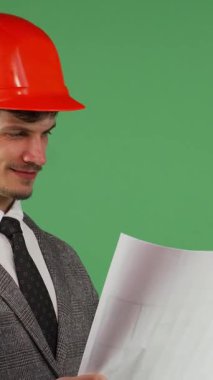Çarpıcı turuncu bir şapka ve şık bir takım elbise giyen neşeli genç bir mimar karmaşık planlarını incelerken gururla ışıldıyor. Canlı yeşil bir krom arka plana yaslanmış, kendine güveni ve ışıl ışıl gülümsemesi...
