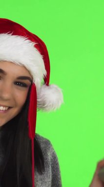 Neşeli, genç bir kadın, neşeli bir Noel Baba şapkası takıyor, güzel paketlenmiş bir Noel hediyesini parlak yeşil renkli bir arka plana karşı tutarken sevinçle parıldıyor. Işıl ışıl gülüşü ve ışıl ışıl gözleri...