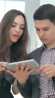 Heyecanlı genç bir çift, şık takım elbiseli bir adam ve şık kıyafetli bir kadın, etkileyici iş sonuçlarını bir tablet üzerinde hevesle analiz ediyorlar. Neşeli ifadeleri ve dinamik takım çalışmaları modern ofisi aydınlatıyor,