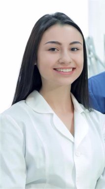 Zarif ve modern bir dişçi ofisinde neşeli bir kadın stajyer ve kendinden emin bir erkek dişçi yan yana durarak sıcaklık ve profesyonellik saçıyor. Baş parmakları ve parlak gülümsemeleri hasta bakımına olan bağlılıklarını yansıtıyor.