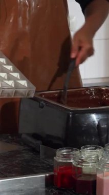 Kendini adamış, parlak bir önlüğe sarılı çikolata, parlak siyah bir kâse içinde kadife erimiş çikolata karıştırır, canlı kalıplar ve renkli lezzet malzemeleriyle çevrilidir. Bu büyüleyici sahne titiz sanatçılık ve tutkuyu vurguluyor.