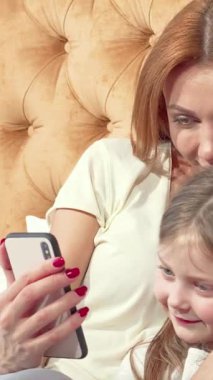 Işıl ışıl bir anne ve sevimli küçük kızı, rahat, altın renkli bir yatağa kurulmuş bir akıllı telefonu keşfederken keyifli gülümsemelerini paylaşıyorlar. Evlerinin sıcak, davetkar atmosferi bağlarını güçlendiriyor, bu da onları rahatlatıyor.