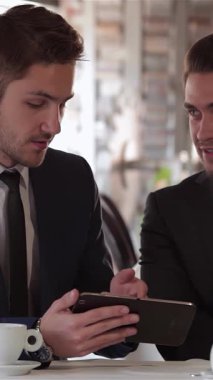 Şık bir kafede, iki iyi giyimli adam hareketli bir restoranın canlı atmosferiyle çevrili bir tableti incelerken yoğun bir şekilde stratejileri tartışıyorlar. Kararlı ifadeleri ve cilalı kıyafetleri profesyonelliği ve işbirliğini simgeliyor.