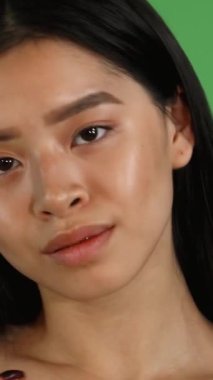 Çekici genç bir Asyalı kadın kameraya güvenle bakarken kusursuz tenini nazikçe okşar. Işıl ışıl güzelliği ve gençlik cazibesi canlı yeşil renkli arka plan tarafından vurgulanıyor, zarafet ve canlılık atmosferi yaratıyor.