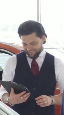 Şık bir donanma yeleği ve kıtır kıtır beyaz gömlek giyen karizmatik bir erkek araba satıcısı, parlak ve modern bir bayilikte potansiyel bir alıcıyla araç seçeneklerini hevesle tartışıyor. Gösterişli otomobiller sergi ışıkları altında parıldıyor.