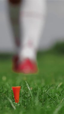 Kırmızıya vuran zarif bir bayan golfçü titizlikle parlak turuncu bir tişörte el değmemiş bir beyaz golf topunu yerleştirir. Odaklanma ve kararlılık yayar. Bu yakın çekim onun eldivenli elini topun üzerinde süzülürken yakalıyor, canlı bir şekilde çerçevelenmiş.