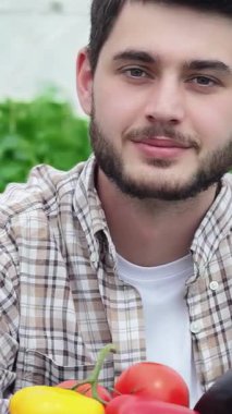 A cheerful young male agronomist beams with pride, presenting a colorful array of freshly harvested organic vegetables, including bright yellow peppers and luscious red tomatoes. This lively scene encapsulates the joy of sustainable farming