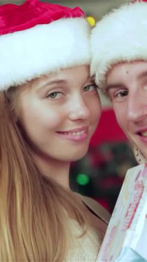 Noel Baba şapkaları ve rahat kazaklar giyen neşeli bir çift, sıcacık aydınlatılmış bir Noel odasında birbirlerine güzel paketlenmiş hediyeler verirken mutluluk saçıyor. Parlak ışıklarla ve canlı bir Noel ağacıyla çevrili, şefkatli gülümsemeleri