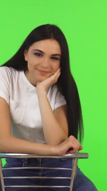 Uzun, dalgalı saçları olan ışıl ışıl genç bir kız modern bir sandalyede zarif bir şekilde oturuyor ve sıcak gülümsemesi sahneyi aydınlatıyor. Çarpıcı yeşil arka plan onun neşeli ruhunu güçlendiriyor, enerjik ve davetkar bir atmosfer yaratıyor yaşam tarzı ve