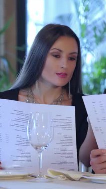 Aşk ve heyecan saçan genç bir çift, yumuşak mum ışığı altında, yemyeşil ile çevrili zarif restoran menüsünü hevesle inceliyor. Neşeli gülümsemeleri ve samimi bakışları büyülü bir ortam yaratır. Kutlamak için mükemmel bir ortam.