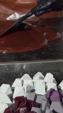 Kendini adamış, siyah eldivenlere bürünmüş, titizlikle yumuşatılmış, cilalı mermer bir yüzey üzerinde erimiş çikolata, renkli, geometrik çikolata kalıplarıyla çevrili. Bu canlı sahne titiz sanatçılık ve tutkuyu yakalıyor.