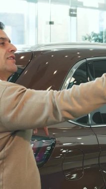 Siyah saçlı şık bir adam şık, lüks bir arabanın önünde, neşe ve güven saçan bir selfie çekerken ışıl ışıl gülümsüyor. Bej renkli bir kazak ve açık beyaz bir gömlek giyiyordu, modern zarafeti somutlaştırıyordu, araçları mükemmel bir şekilde tamamlıyordu.