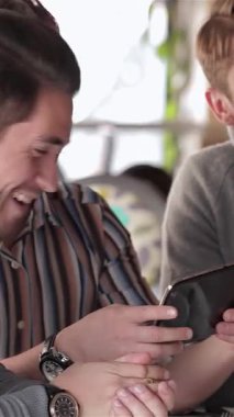 Canlı bir kafe ortamında, rahat giyimli üç genç işadamı neşeli bir tartışmaya dalmış, yüzleri neşe ve dostlukla aydınlanıyordu. Sıcak atmosfer, hareketleriyle ve ortak gülüşleriyle zenginleşir.