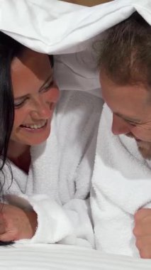 Pofuduk beyaz bir battaniyeye sarılmış neşeli olgun bir çift, yatakta yatarken bulaşıcı gülüşler ve sevgi dolu bakışlar paylaşıyor. Işıl ışıl gülüşleri ve neşeli ifadeleri samimi, samimi bir atmosfer yaratıyor. Aşkı kutlamak için mükemmel.