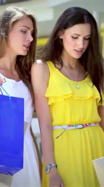 İki şık kız, biri canlı sarı elbiseli, diğeri şık beyaz elbiseli, renkli alışveriş torbalarını gururla sergiliyorlar, neşe ve dostluk saçıyorlar. Işıldayan gülümsemeleri ve canlı atmosferleri gençliğin coşkusunu yansıtıyor.