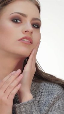 Uzun, dalgalı esmer saçlı neşeli genç bir kadın kameraya güvenli bir şekilde poz veriyor, ışıl ışıl gülüşü el değmemiş beyaz bir fonda sahneyi aydınlatıyor. Gri bir süveter giyerek modern zarafet ve gençlik cazibesi yayıyor.