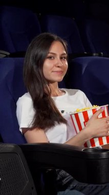 Uzun, dalgalı saçları olan çekici genç bir kadın rahat bir şekilde rahat bir sinema koltuğunda oturur, canlı çizgili patlamış mısır kovasını sallarken yanındaki boş koltuğu neşeyle işaret eder. Işıltılı gülüşü ve gevşemiş tavırları davetkar bir etki yaratıyor.