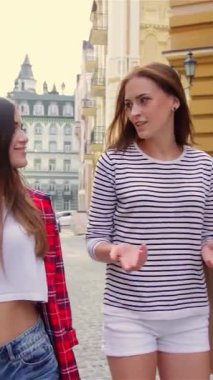 İki neşeli genç kadın, ellerinde hoş bulgularla dolu renkli alışveriş torbaları tutarak, gülerek ve hikayeler paylaşarak, pitoresk bir şehir caddesinde yürüyorlar. Şık kıyafetleri ve ışıl ışıl gülümsemeleri yazın kaygısız ruhunu temsil ediyor.