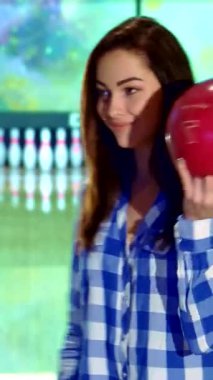 Işıl ışıl bir bowling salonunun ışıl ışıl şeritlerine çarpan kırmızı bowling topunu sallarken kendine güvenen esmer saçlı çekici beyaz bir kız. Şık, kareli bir gömlek giymiş, neşeli duruşu canlandırıcı.