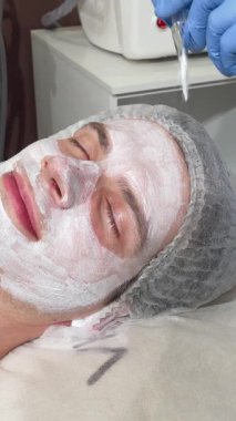 Huzurlu bir adam güzellik salonunda uzanır, yetenekli bir güzellik uzmanının titizlikle kremli bir yüz maskesi takması gibi gözleri mutlulukla kaplıdır. Bu rahatlatıcı tedavi, kendine bakmanın ve rahatlamanın özünü somutlaştırıyor.