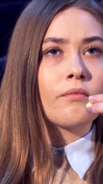 Uzun, dalgalı kahverengi saçlı beyaz bir kızın büyüleyici yakın çekimi, yoğun bakışları kendisini sürükleyici bir filme daldırırken derin bir endişeyi ortaya koyuyor. Loş ışıklı tiyatroda, geniş gözleri ekrandaki dramı yansıtırken, gergin bir şekilde