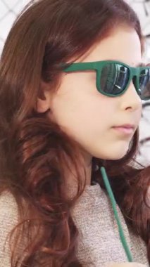 Kestane rengi saçları akan tatlı küçük bir kız neşeli bir gözlük mağazasında neşe saçan ve kendine güvenen şık yeşil güneş gözlüğü deniyor. Renkli çerçevelerin kaleydoskopu ile çevrelenmiş, çocukluk modasının özünü yakalıyor.