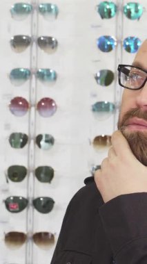 Düşüncelere dalmış, olgun sakallı bir adamın yakın çekimi. Bir gözlük mağazasında sergilenen göz kamaştırıcı gözlük dizisini incelerken sakalını nazikçe okşuyor. Bu büyüleyici sahne, kişisel stil ve tüketici seçiminin yolculuğunu somutlaştırıyor.