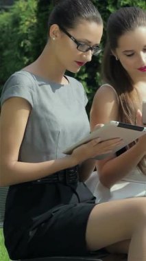 Canlı bir şehir parkında, iki şık iş kadını tabletlerine derinden odaklanırken, şık takım elbiseli üçüncü bir kadın da yakınlarda önemli bir telefon alıyor. Bu canlı sahne modern kurumsal hayatın nabzını somutlaştırıyor.