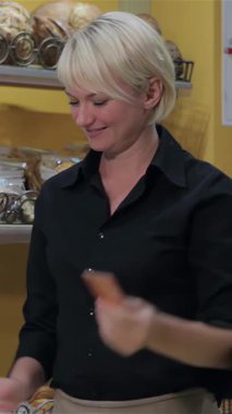 Renkli bir pastanede müşterilerinin kredi kartı ödemelerini işlerken şık siyah gömlekli ve önlüklü hoş bir genç sarışın kadın. Baştan çıkarıcı yeni pişmiş mallar, sıcak gülümsemesi ve çekici tavırları ile çevrili.