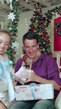 Keyifli bir çift, rahat kazaklara ve canlı eşarplara sarılmış, harika dekore edilmiş bir Noel odasında güzel paketlenmiş hediyeleri hevesle açıyor. Parlayan ışıklarla çevrili ve görkemli bir Noel ağacı ile süslenmiş,