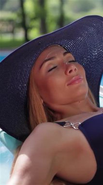 Güzel genç bir kadın, sıcak güneş ışığında ışıldayan yüzme havuzunda zarifçe uzanır. Şık, koyu mavi bir mayo ve şık, geniş kenarlı bir mavi şapkayla süslenip kollarını başının üstüne uzatarak rahatlıyor.