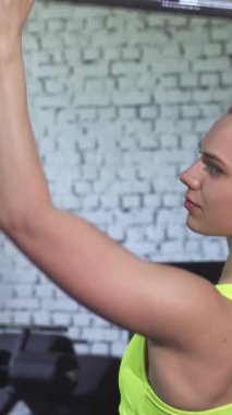 Uzun saçlı odaklanmış genç bir kadın, son bir çekme egzersizi yaparken kendine güven yayıyor, çarpıcı neon bir spor bluzu içinde kollarını esnetiyor. Parlak ışıklarla aydınlatılan gösterişli jimnastik salonu gücünü ve kararlılığını arttırıyor.