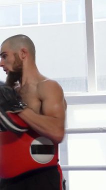 Kararlı üstsüz bir erkek kickboksör, odaklanmış koçuyla antrenman yaparken güçlü tekmeler ve yumruklar atıyor. O da doğal ışıkla dolu canlı bir spor salonunda kırmızı pedleri ustalıkla tutuyor. Atmosfer hem yoğunluk hem de motivasyon ile uğulduyor.