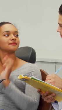 Dost canlısı bir erkek dişçi, güzel Afrikalı kadın hastasıyla sıcak bir sohbet için tıbbi evrakları dikkatlice doldurur. Bu davet sahnesi, iletişimin ve diş bakımına duyulan güvenin önemini vurguluyor.