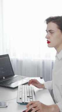 Gıcır gıcır beyaz bir gömlek giyen yorgun genç bir kadın, dizüstü bilgisayarı ve masaüstündeki işlerle uğraşırken gözle görülür bir şekilde şaşkına dönmüş bir şekilde modern ofis masasında oturuyor. Perdelerin arasından süzülen yumuşak, doğal ışık onun stresli ifadesini vurguluyor.