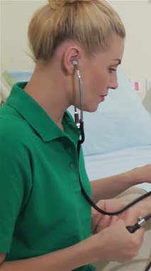 Canlı yeşil polo giymiş, kendini adamış genç bir beyaz hemşire, rahat bir hastane yatağında dinlenen yaşlı bir adamın kan basıncını dikkatle ölçüyor. Yumuşak, sıcak aydınlatmayla aydınlatılan sakin ortam, onun profesyonelliğini vurguluyor.