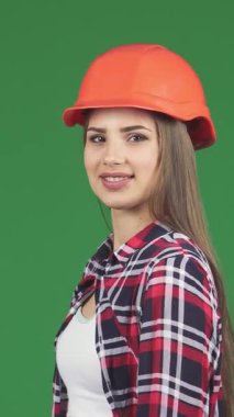 Çarpıcı genç bir kadın yapıcı, canlı turuncu bir şapka ve şık bir ekose gömlek ile süslenmiş, canlı yeşil kromakey arka planına karşı kaslarını güvenli bir şekilde esnetir. Bu güç verici görüntü güç ve feminizm yayıyor.