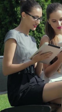 Canlı bir açık havada, şık giyinmiş iki şık iş kadını, bir tablet üzerinde özgüven ve profesyonellik yayarak birlikte çalışırlar. Bu arada, arka planda üçüncü bir kadın bir telefon görüşmesi yapıyor.