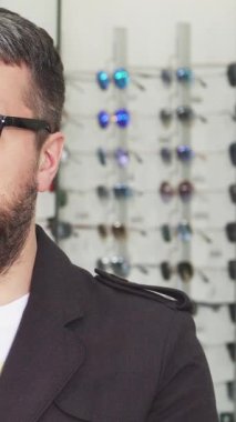 Sakallı bir adam şık bir optometrist dükkanında elinde iki adet şık güneş gözlüğü ağırlığında duruyor. Göz kamaştırıcı renkli kıyafetlerle çevrelenmiş, modern tüketiciliğin bir örneği, stil ve moda için ayırt edici bir tat sergiliyor.