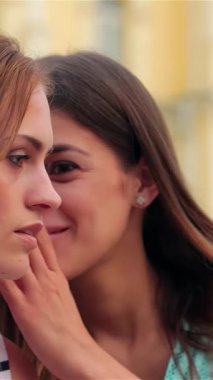 İki güzel kız birbirine yakın bir şekilde eğiliyor, hayat dolu bir kasaba meydanında sırlarını fısıldıyor, neşe ve dostluk saçıyor. Işıl ışıl gülüşleri ve günlük yaz kıyafetleri genç arkadaşlığın özünü yansıtıyor.