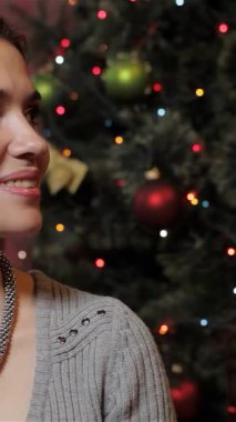 Keyifli bir esmer kadın, hoş kulaklıklar takıyor, göz kamaştırıcı bir Noel ağacının arka planına başparmak sallıyor. Parıldayan ışıklar ve canlı süsler sıcak, şenlikli bir ortam yaratarak herkesi bayramı paylaşmaya davet ediyor.