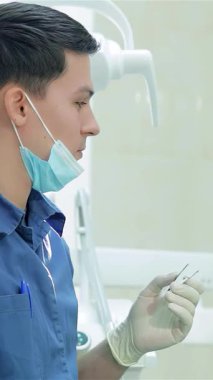 Mavi üniformalı ve maskeli gülümseyen bir dişçi, dişçi aletlerini dikkatlice seçer, hastasına özen ve bakım sağlar. Parlak, steril klinik profesyonelliği yayıyor, rahat bir diş sağlığı deneyimi için hoş bir ortam yaratıyor.