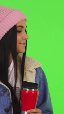 Hoş bir genç kadın, şık bir kot ceket ve yumuşak pembe bir bere ile kaplanmış, sıcak çayının tadını çıkarıyor, canlı yeşil renkli bir arka plana sıcak bir ışıltı yayıyor. Bu rahat sahne konfor ve mutluluğun özünü özetliyor.