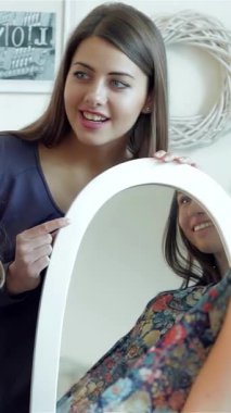 İki modaya uygun kız arkadaş, şık bir elbiseyi parlak bir aynanın önünde heyecanlı bir şekilde ölçerken, yüzleri neşe ve heyecanla parlarken aynı anı paylaşıyorlar. Bu canlı sahne, arkadaşlığın ve modanın heyecanını yansıtıyor.