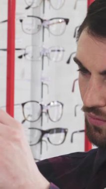 Yakışıklı, genç, sakallı bir adam yakın plan, keskin bir bakışla, canlı bir göz teçhizatı arasında parlak bir çift gözlük teftiş. Arka plandaki renkli çerçeveler canlı bir alışveriş ortamı yaratıyor, modayla işlevselliği harmanlıyor.